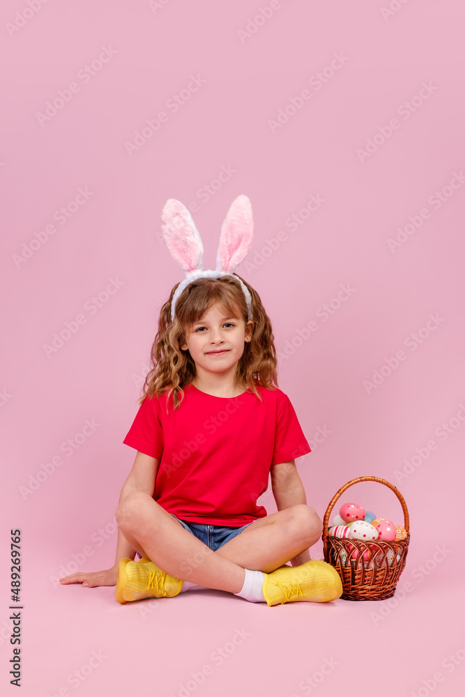 Little beautiful smiling girl in Easter bunny ears headband sitting on the floor near basket with Easter eggs on pink background. Copy space, vertical orientation