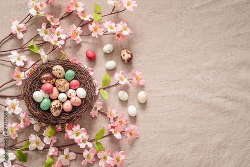 Easter background with bird’s nest, colored easter eggs and blooming brunches on a beige background. Easter composition on a textured beige linen background. 