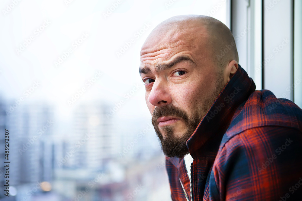 Portrait of an attractive bald man with a beard in casual clothes at the window, he looks into the camera with a frown, puzzled and displeased. Outside the window is a blurry landscape of a big city. Portrait of an attractive bald man with a beard in casual clothes at the window, he looks into the camera with a frown, puzzled and displeased. Outside the window is a blurry landscape of a big city.