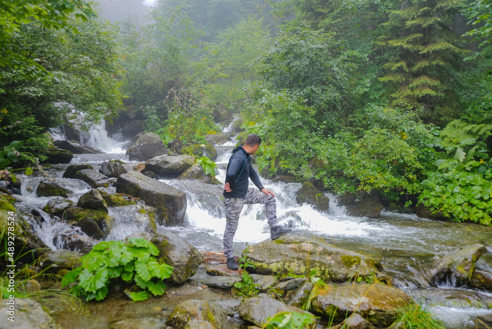 A young man admires the beauty of the waterfall. A beautiful view in the forest with a river with a small waterfall among mountain rocks, with a small breeze of fog. nature's beauty