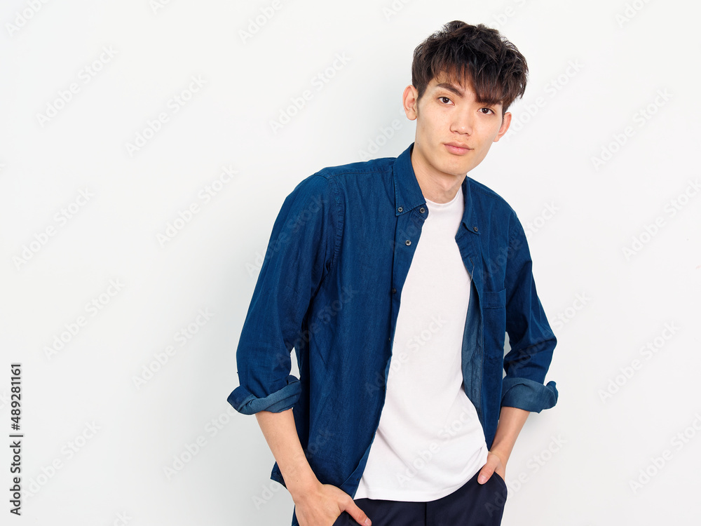 Portrait of handsome Chinese young man with curly black hair in blue shirt posing against white wall background. Hands in his pockets and smiling at camera, looks confident, front view. Portrait of handsome Chinese young man with curly black hair in blue shirt posing against white wall background. Hands in his pockets and smiling at camera, looks confident, front view.