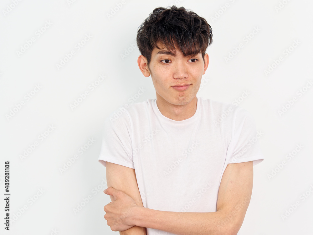 Portrait of handsome Chinese young man with curly black hair in white T-shirt posing against white wall background. Hand on arm and looking away with funny expression, looks frustrated, front view. Portrait of handsome Chinese young man with curly black hair in white T-shirt posing against white wall background. Hand on arm and looking away with funny expression, looks frustrated, front view.