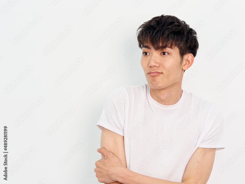 Portrait of handsome Chinese young man with curly black hair in white T-shirt posing against white wall background. Hand on arm and looking away with funny expression, looks frustrated, front view. Portrait of handsome Chinese young man with curly black hair in white T-shirt posing against white wall background. Hand on arm and looking away with funny expression, looks frustrated, front view.