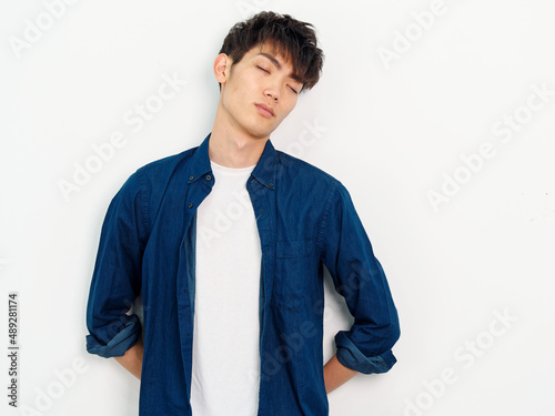 Portrait of handsome Chinese young man with curly black hair in blue shirt posing against white wall background. Hands on his back and eyes closed with tired expression, looks exhausted.