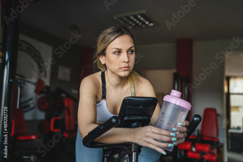 One woman at the gym training hold supplement shaker while riding spinning bike during workout real people caucasian female endurance and healthy life concept supplementation drink copy space