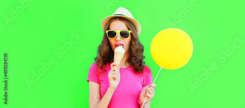 Portrait of young woman eating ice cream with balloon wearing summer straw hat on green background