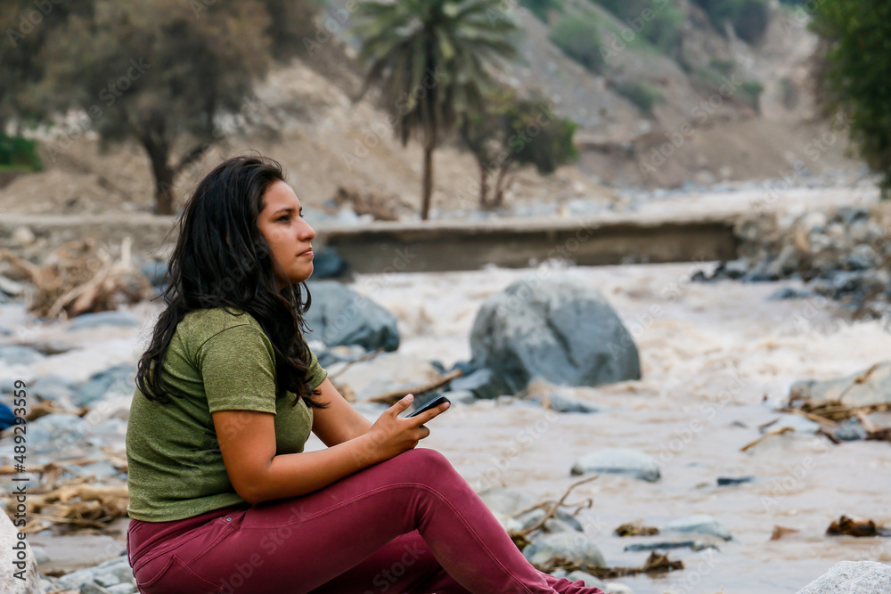 Woman sitting on a river bank holding a cell phone with her hands.