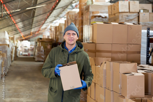 Caucasian man worker holding cardboard box while standing in storehouse, looking at camera and smiling.