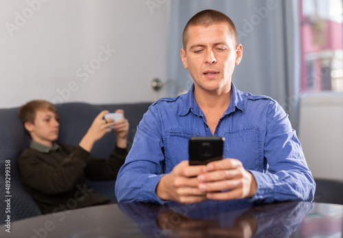 Positive american man sitting at kitchen with smartphone and looking at screen, his son sitting at sofa