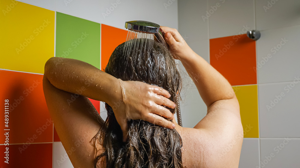 Naklejka premium Woman rinsing shampoo from her long dark hair with a jet of water from the shower. Back view. Multicolored tiles in the background. The concept of hair care and personal hygiene.