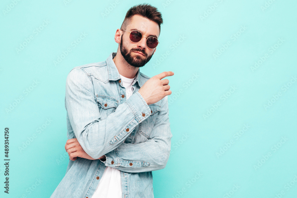Portrait of handsome confident stylish hipster lambersexual model.Man dressed in jacket and jeans. Fashion male posing near blue wall in studio in sunglasses Portrait of handsome confident stylish hipster lambersexual model.Man dressed in jacket and jeans. Fashion male posing near blue wall in studio in sunglasses