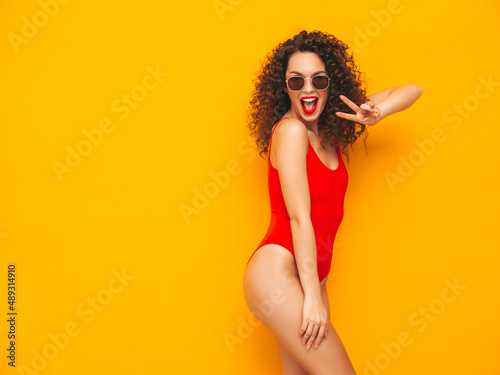 Young beautiful smiling woman posing near yellow wall in studio.Sexy model in red swimwear bathing suit.Positive female with curls hairstyle.Happy and cheerful. In sunglasses © halayalex
