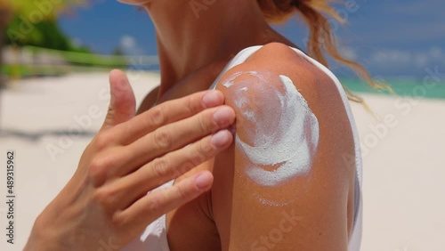 Close up of a happy smiling young woman is applying a sunscreen or sun tanning lotion on a shoulder to take care of her skin on a seaside beach during holidays vacation.