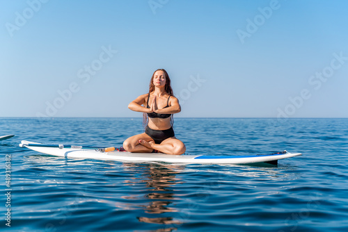 Sporty girl on a glanders surfboard in the sea on a sunny summer day. In a striped swimsuit, he sits in the lotus position with his eyes closed.