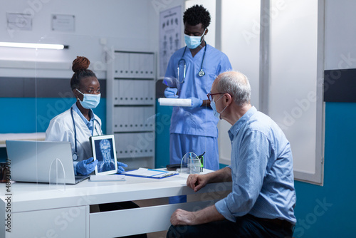 Clinic radiology specialist holding x-ray scan image and explaining illness to retired man while medical nurse taking notes on clipboard. Expert radiologist showing MRI scan image to elderly man.