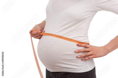 Closeup of Hands of Pregnant Caucasian Woman Posing With Measurement Tape In White Shirt While Taking Dimensions of Her Belly On White Background. photo