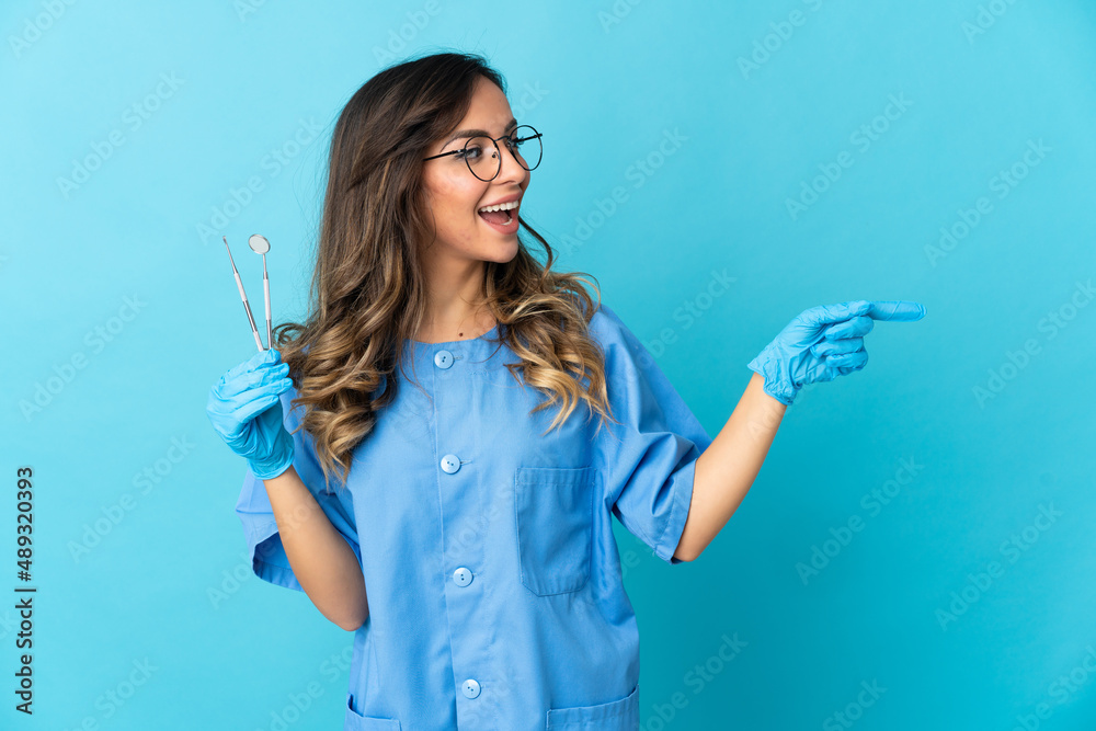 Woman dentist holding tools over isolated on blue background pointing finger to the side and presenting a product Woman dentist holding tools over isolated on blue background pointing finger to the side and presenting a product