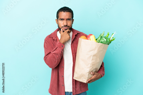Young latin man holding a grocery shopping bag isolated on blue background thinking