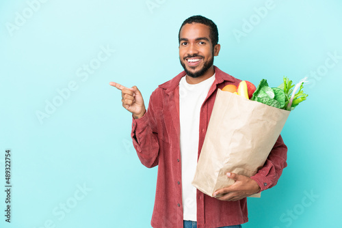 Young latin man holding a grocery shopping bag isolated on blue background pointing finger to the side