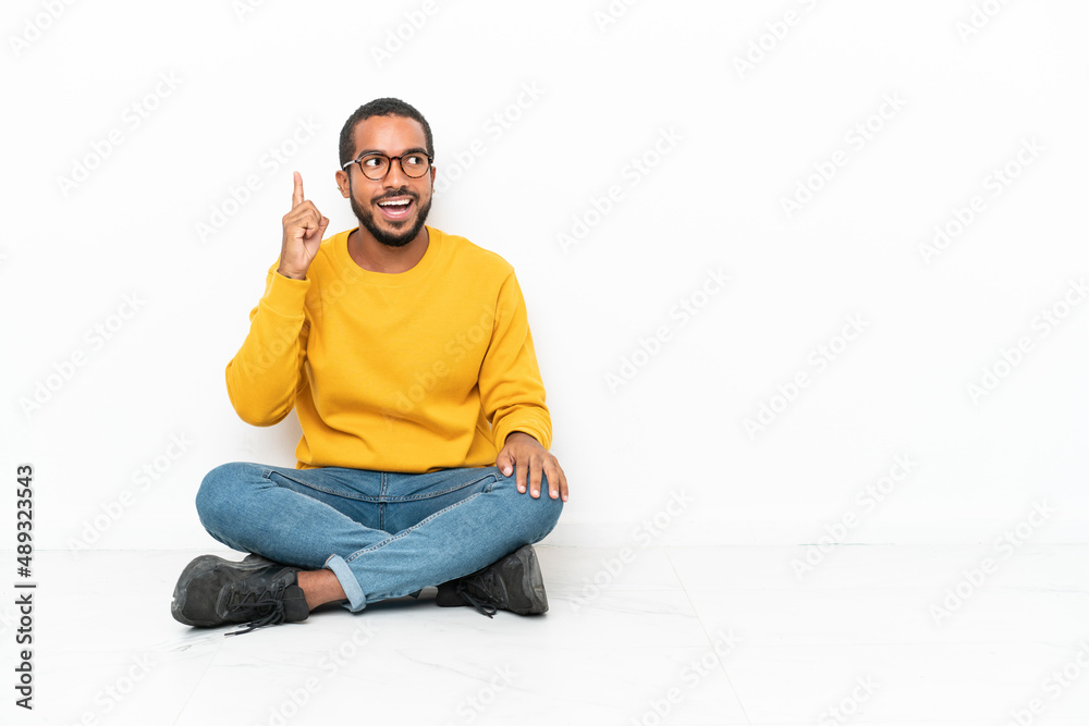 Young Ecuadorian man sitting on the floor isolated on white wall intending to realizes the solution while lifting a finger up Young Ecuadorian man sitting on the floor isolated on white wall intending to realizes the solution while lifting a finger up