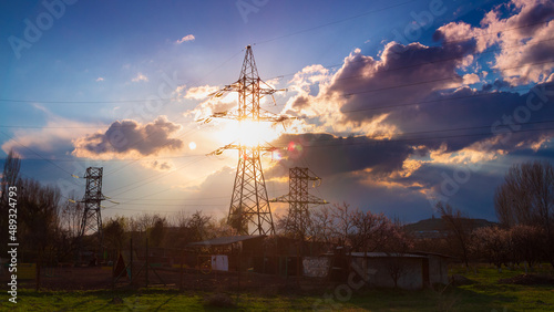 High voltage electrical pole structure on background dramatic sunset with fluffy clouds, golden sun, trees and blue sky
