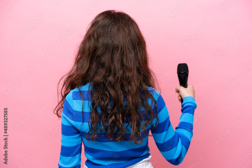 Little singer girl picking up a microphone isolated on pink background in back position Little singer girl picking up a microphone isolated on pink background in back position