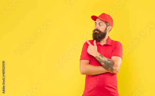 Serious deliveryman in casual red cap and tshirt pointing finger aside copy space  advert