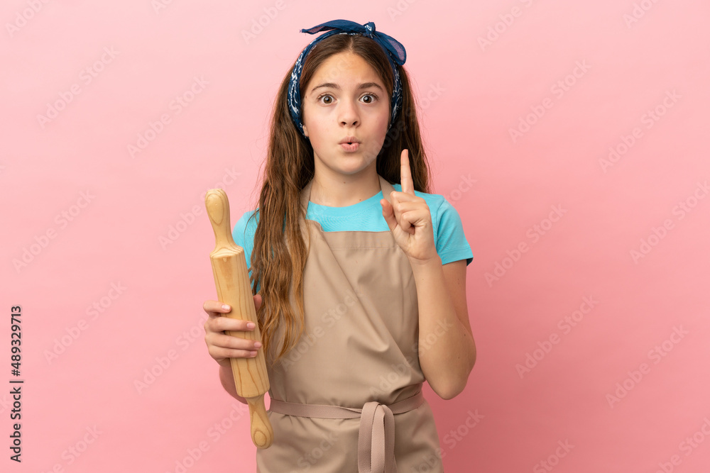 Little caucasian girl holding a rolling pin isolated on pink background intending to realizes the solution while lifting a finger up Little caucasian girl holding a rolling pin isolated on pink background intending to realizes the solution while lifting a finger up