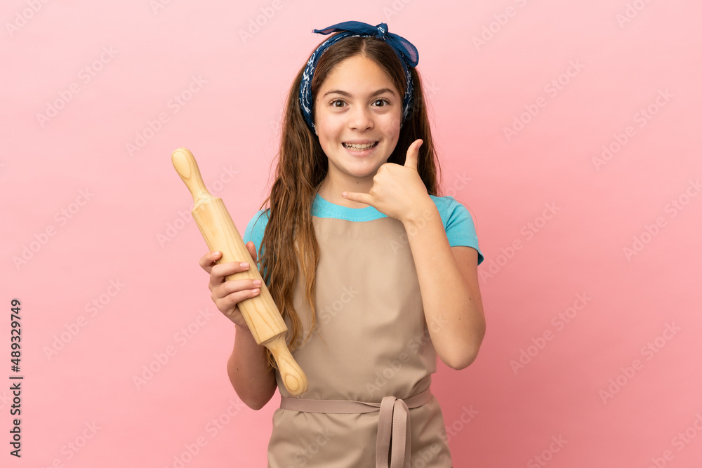 Little caucasian girl holding a rolling pin isolated on pink background making phone gesture. Call me back sign Little caucasian girl holding a rolling pin isolated on pink background making phone gesture. Call me back sign