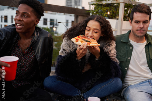 Group of multi-cultural friends sitting eating delicious pizza on a rooftop terrace. Biracial female biting into food.