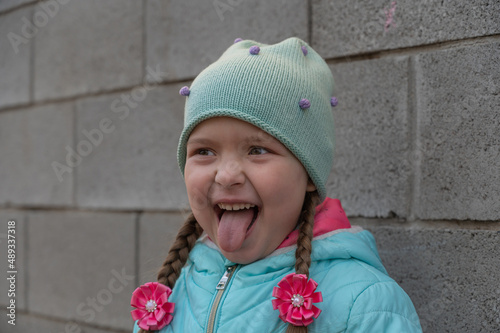 The granddaughter grimaces against the gray wall. A five-year-old child with long hair braided into pigtails. A preschooler in a blue jacket and hat shows his tongue and laughs. Springtime.
