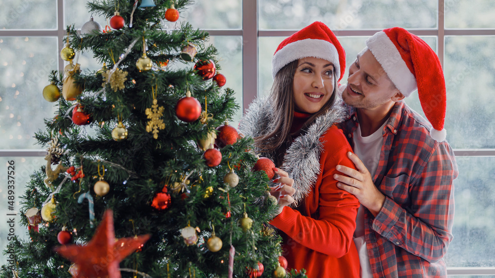 Caucasian young happy lover couple husband and wife in red Santa Claus hat and sweater holding helping each other hanging ornamental bauble sphere ball on Xmas tree in living room in Christmas eve Caucasian young happy lover couple husband and wife in red Santa Claus hat and sweater holding helping each other hanging ornamental bauble sphere ball on Xmas tree in living room in Christmas eve