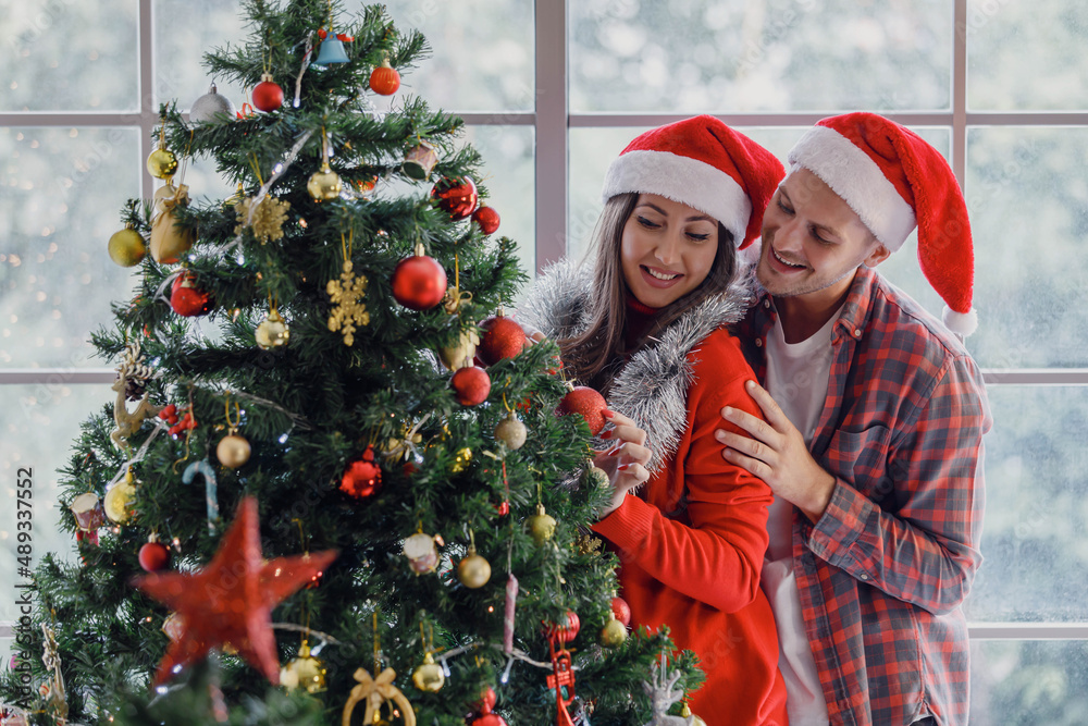 Caucasian young happy lover couple husband and wife in red Santa Claus hat and sweater holding helping each other hanging ornamental bauble sphere ball on Xmas tree in living room in Christmas eve Caucasian young happy lover couple husband and wife in red Santa Claus hat and sweater holding helping each other hanging ornamental bauble sphere ball on Xmas tree in living room in Christmas eve