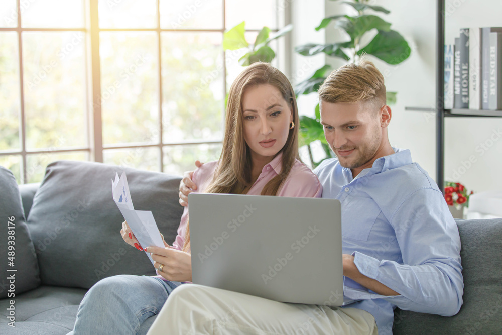 Millennial caucasian lover couple husband and wife in casual outfit sitting smiling on sofa surfing browsing internet shopping online together via laptop notebook computer in living room on weekend Millennial caucasian lover couple husband and wife in casual outfit sitting smiling on sofa surfing browsing internet shopping online together via laptop notebook computer in living room on weekend