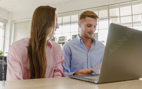 Millennial caucasian lover couple husband and wife in casual outfit sitting smiling on sofa surfing browsing internet shopping online together via laptop notebook computer in living room on weekend