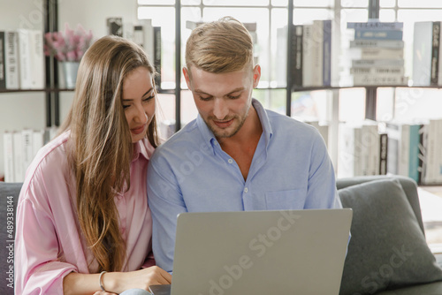 Millennial caucasian lover couple husband and wife in casual outfit sitting smiling on sofa surfing browsing internet shopping online together via laptop notebook computer in living room on weekend