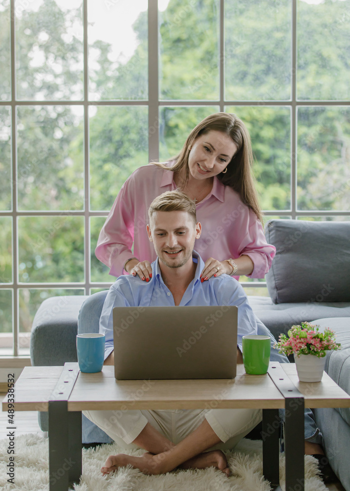 Millennial young Caucasian lover couple handsome husband sitting on floor browsing surfing internet shopping online via laptop computer while beautiful wife on cozy sofa help massaging shoulders. Millennial young Caucasian lover couple handsome husband sitting on floor browsing surfing internet shopping online via laptop computer while beautiful wife on cozy sofa help massaging shoulders.