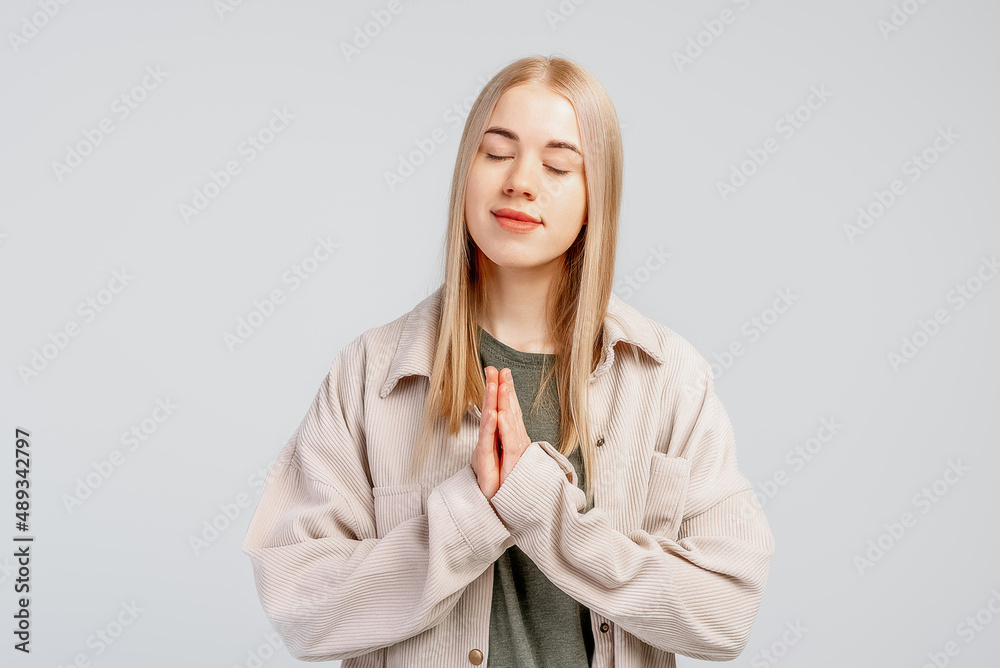 Peaceful and mindful fair-haired young woman praying, holding hands in pray, namaste gesture and close eyes, relaxing and breathing freely, standing over grey background