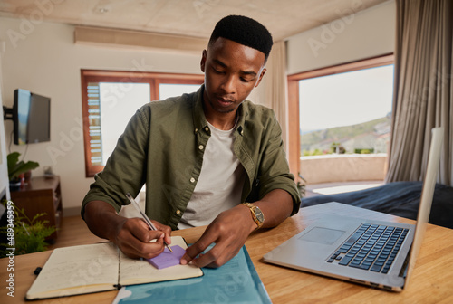 Young adult black male writing note on sticky note next to laptop, concentrating. Sitting in modern apartment working from home.