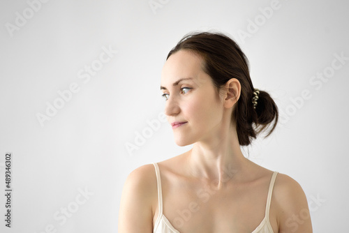 authentic portrait of brunette woman with gathered hair in beige T-shirt with look to side on white background  tired mother without makeup