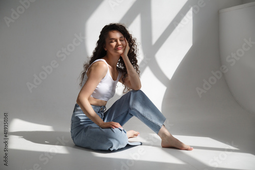 A cute slender girl with long curly hair sits at a bright room on a sunny day.