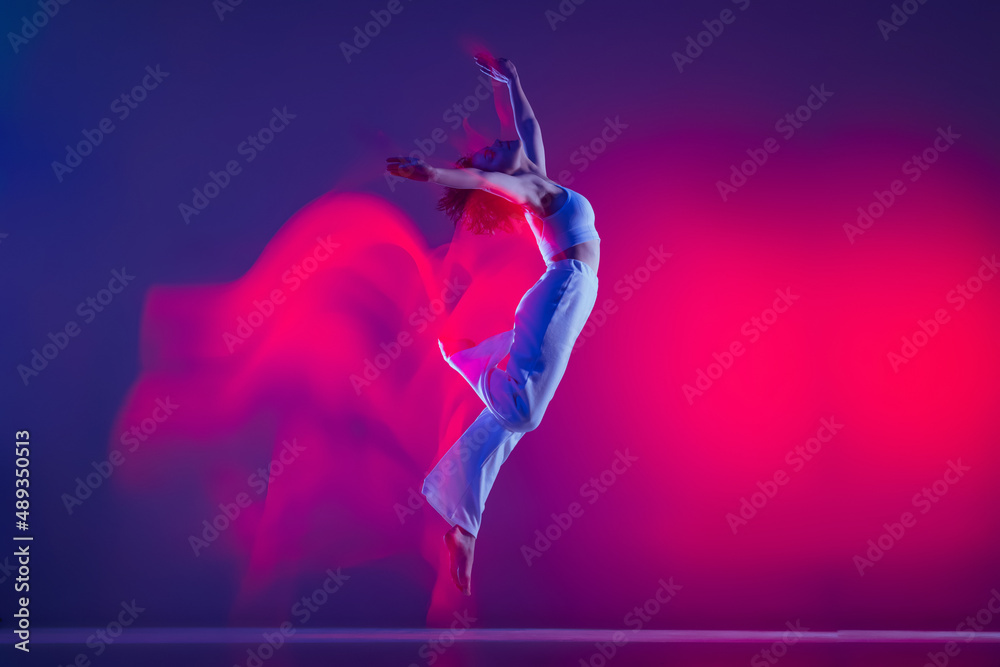 Flight. Young sportive beautiful girl, hip-hop dancer dancing hip hop isolated on purple background in pink neon light. Youth culture, style and fashion, action.