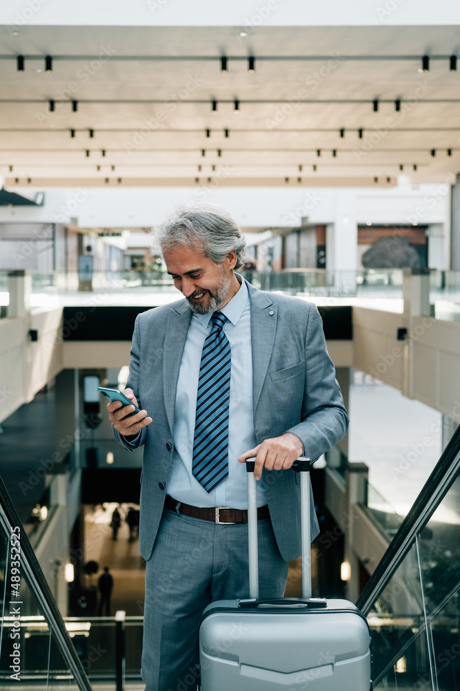 Smiling Mature Businessman Standing on Escalator and Using his Mobile Phone at the Airport Smiling Mature Businessman Standing on Escalator and Using his Mobile Phone at the Airport
