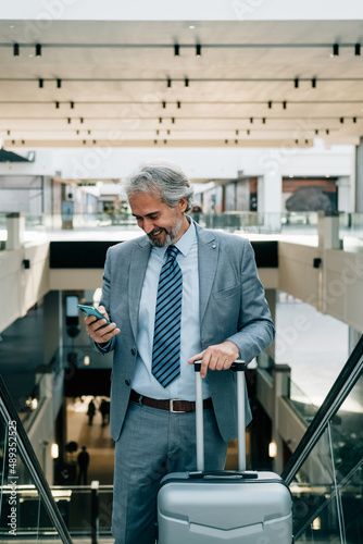 Smiling Mature Businessman Standing on Escalator and Using his Mobile Phone at the Airport