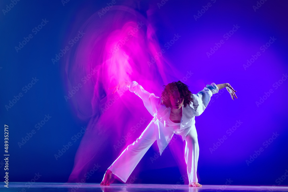 Dynamic portrait of beautiful girl, hip-hop dancer in white outfit dancing hip hop isolated on blue background in pink neon light. Youth culture, style and fashion, action.