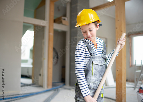 cool boy with yellow safety helmet and dungarees on construction site in a house and loft with shovel in hand