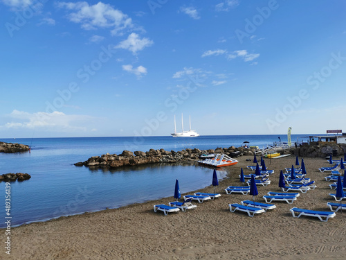 Sandy beach in a bay with sun loungers and umbrellas, in the Mediterranean Sea the largest sailing yacht in the world, an eight-deck motorsailer against a blue sky with clouds. © Elena