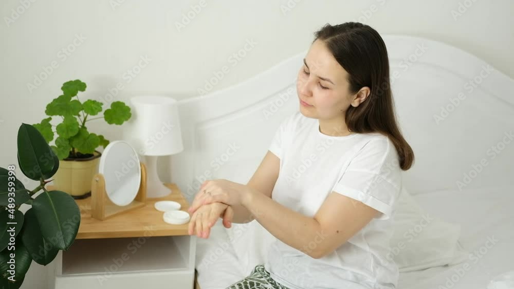 Young caucasian woman, brunette girl, is sitting on bed and smearing her hands with moisturizer. Home cosmetic spa care, hand massage. Young caucasian woman, brunette girl, is sitting on bed and smearing her hands with moisturizer. Home cosmetic spa care, hand massage.