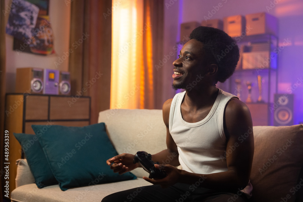 A smiling cheerful dark-skinned man sits on a couch in the living room holding a game pad from a console in his hands, looking at the screen satisfied