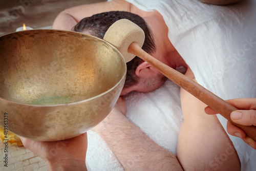 A woman performs Tibetan singing bowl therapy with a man lying under a white sheet. Relaxing meditation photo